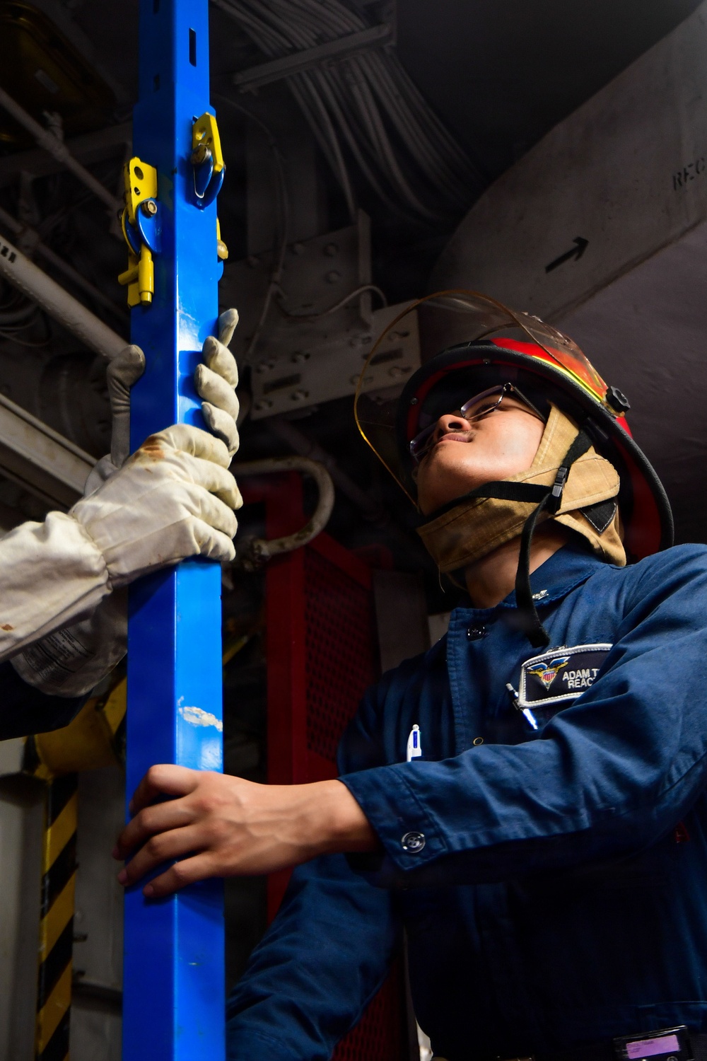 A person wearing a firefighter/damage control helmet with a face shield, safety glasses, and work gloves is handling a blue vertical support pole or shoring device in what appears to be a naval vessel interior. The individual is dressed in a blue coverall uniform with a name tag partially visible reading 'ADAM T.' and 'REAC...' suggesting a damage control or emergency response role. The setting appears to be a ship's hangar bay or interior compartment with industrial equipment visible in the background.