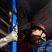 A person wearing a firefighter/damage control helmet with a face shield, safety glasses, and work gloves is handling a blue vertical support pole or shoring device in what appears to be a naval vessel interior. The individual is dressed in a blue coverall uniform with a name tag partially visible reading 'ADAM T.' and 'REAC...' suggesting a damage control or emergency response role. The setting appears to be a ship's hangar bay or interior compartment with industrial equipment visible in the background.