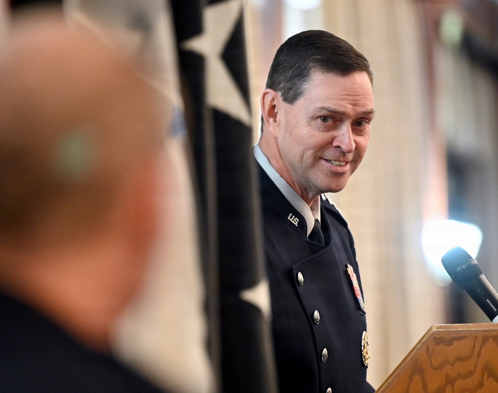A U.S. military officer in dress uniform stands at or near a podium, appearing to speak or listen at a formal ceremony. He is wearing a dark blue dress uniform with 'U.S.' collar insignia, decorations, and brass buttons. A star-bearing flag or banner is visible in the background.