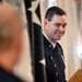 A U.S. military officer in dress uniform stands at or near a podium, appearing to speak or listen at a formal ceremony. He is wearing a dark blue dress uniform with 'U.S.' collar insignia, decorations, and brass buttons. A star-bearing flag or banner is visible in the background.