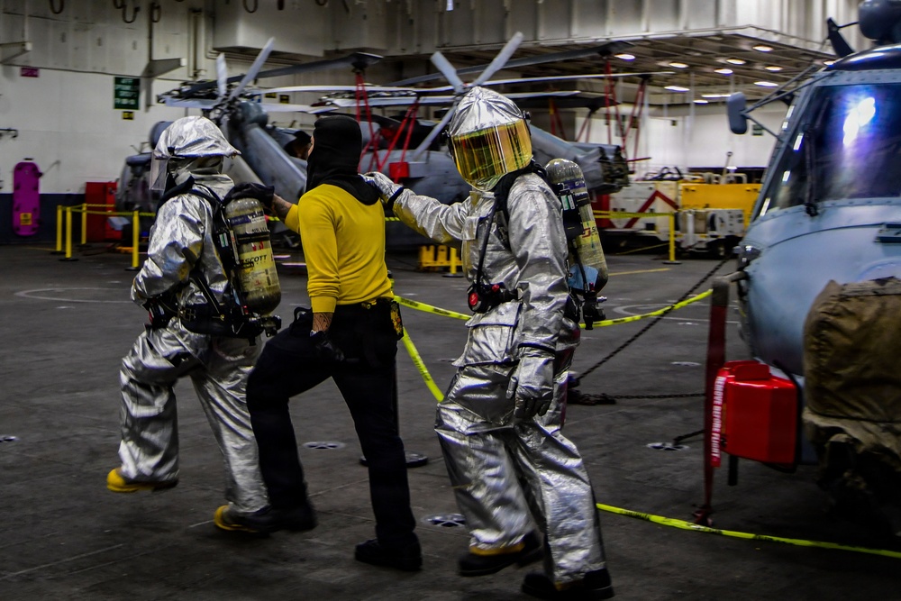The image shows what appears to be a damage control or firefighting drill aboard a naval vessel's hangar deck. Two personnel in silver aluminized proximity/firefighting suits with SCBA (Self-Contained Breathing Apparatus) units are escorting or assisting a third person in a yellow shirt and black balaclava. Military helicopters are visible in the background, confirming the setting as a ship's hangar bay.