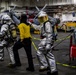 The image shows what appears to be a damage control or firefighting drill aboard a naval vessel's hangar deck. Two personnel in silver aluminized proximity/firefighting suits with SCBA (Self-Contained Breathing Apparatus) units are escorting or assisting a third person in a yellow shirt and black balaclava. Military helicopters are visible in the background, confirming the setting as a ship's hangar bay.