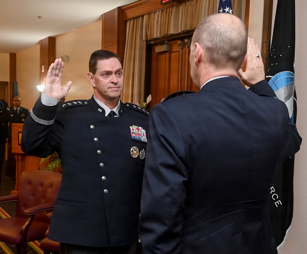 A military swearing-in or oath-taking ceremony is taking place in a formal indoor setting. Two uniformed officers are facing each other with right hands raised, indicating an oath of office or promotion ceremony. A dark flag with what appears to be a space or intelligence agency insignia is visible in the background.