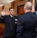A military swearing-in or oath-taking ceremony is taking place in a formal indoor setting. Two uniformed officers are facing each other with right hands raised, indicating an oath of office or promotion ceremony. A dark flag with what appears to be a space or intelligence agency insignia is visible in the background.