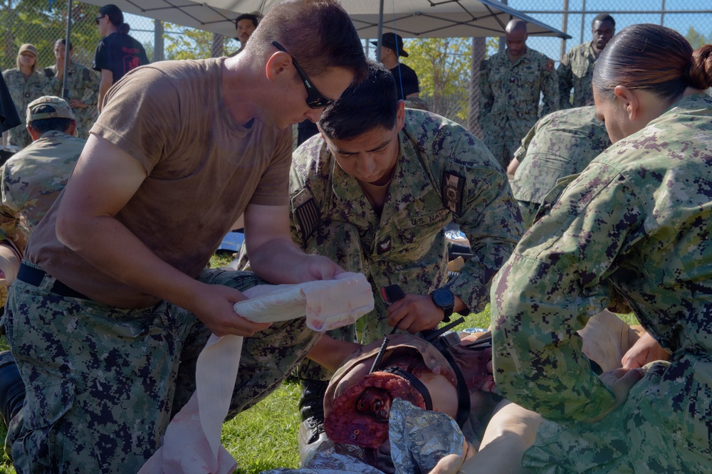 U.S. Navy personnel are conducting a medical training exercise outdoors, practicing trauma care on a simulated casualty with a realistic wound moulage. Three primary participants are actively applying bandaging and wound treatment to a person lying on the ground. The setting appears to be a military training event with multiple uniformed personnel visible in the background.