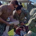 U.S. Navy personnel are conducting a medical training exercise outdoors, practicing trauma care on a simulated casualty with a realistic wound moulage. Three primary participants are actively applying bandaging and wound treatment to a person lying on the ground. The setting appears to be a military training event with multiple uniformed personnel visible in the background.