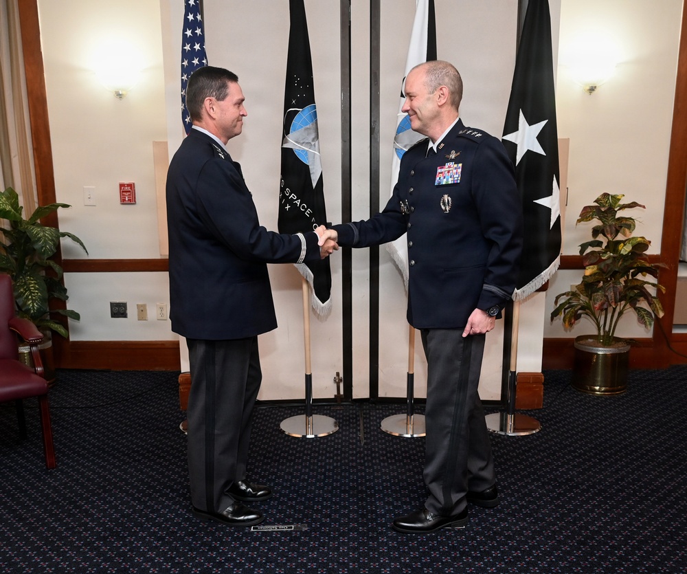 Two military officers in dark dress uniforms are shaking hands in a formal ceremonial setting. The officer on the right displays multiple service ribbons and medals, suggesting a senior rank. Flags including the United States flag and what appears to be a Space Command flag are displayed in the background.