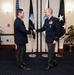 Two military officers in dark dress uniforms are shaking hands in a formal ceremonial setting. The officer on the right displays multiple service ribbons and medals, suggesting a senior rank. Flags including the United States flag and what appears to be a Space Command flag are displayed in the background.