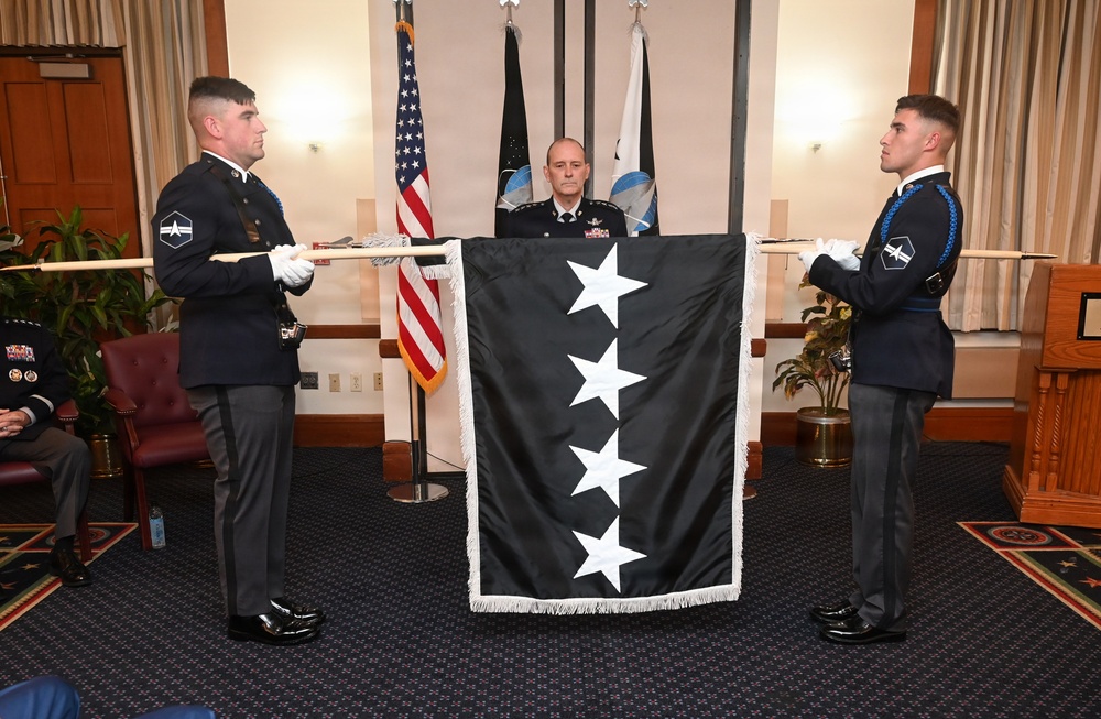 A military ceremony is taking place indoors, featuring two uniformed honor guard members displaying a black flag with four white stars arranged diagonally, consistent with a four-star general's flag. A senior officer in a decorated uniform stands behind the flag, suggesting a promotion or change of command ceremony. The setting appears to be a formal military venue with U.S. and Space Force flags visible in the background.