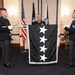 A military ceremony is taking place indoors, featuring two uniformed honor guard members displaying a black flag with four white stars arranged diagonally, consistent with a four-star general's flag. A senior officer in a decorated uniform stands behind the flag, suggesting a promotion or change of command ceremony. The setting appears to be a formal military venue with U.S. and Space Force flags visible in the background.