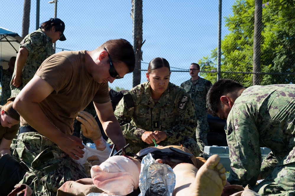 Military personnel in various uniforms are conducting what appears to be a medical training exercise outdoors. A U.S. Navy service member (name tag partially visible as 'HUNT') and others are performing simulated casualty care on a training mannequin or simulated patient. The setting appears to be a fenced outdoor area, likely a military training facility.