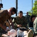 Military personnel in various uniforms are conducting what appears to be a medical training exercise outdoors. A U.S. Navy service member (name tag partially visible as 'HUNT') and others are performing simulated casualty care on a training mannequin or simulated patient. The setting appears to be a fenced outdoor area, likely a military training facility.