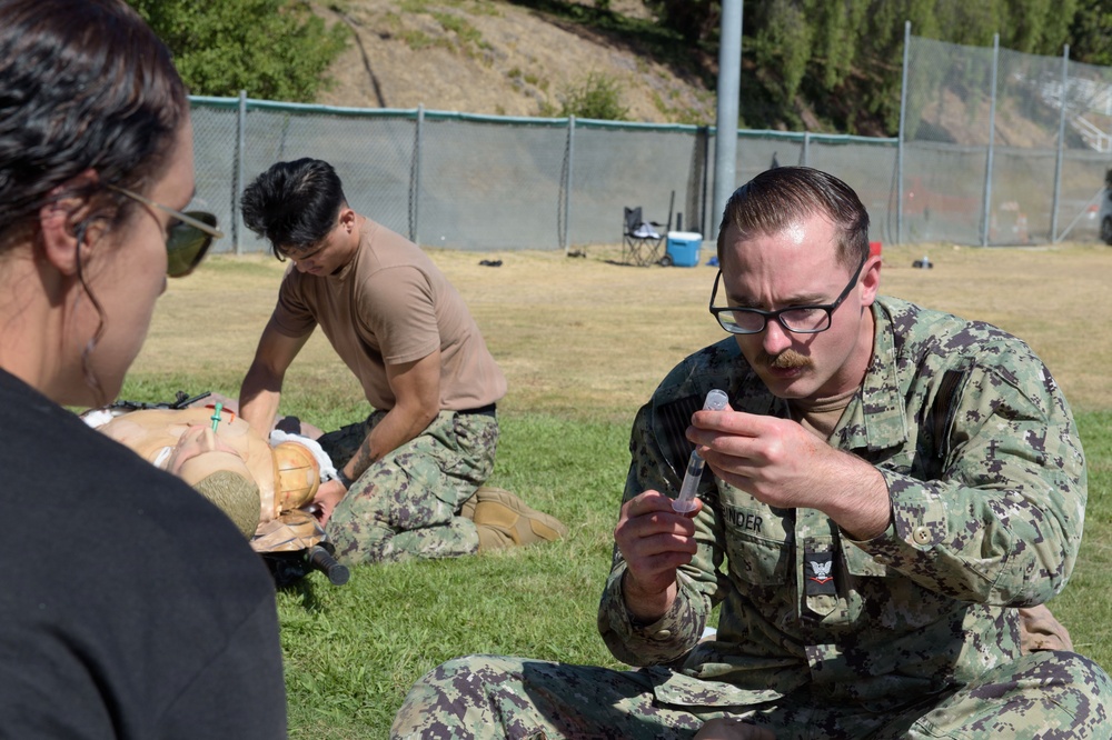 Military personnel in U.S. Navy digital camouflage uniforms are conducting an outdoor medical training exercise on a grass field. One sailor in the foreground is examining a syringe or medical instrument, while another in the background works on a medical training mannequin. A civilian or instructor in a black shirt observes from the left side of the frame.