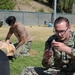 Military personnel in U.S. Navy digital camouflage uniforms are conducting an outdoor medical training exercise on a grass field. One sailor in the foreground is examining a syringe or medical instrument, while another in the background works on a medical training mannequin. A civilian or instructor in a black shirt observes from the left side of the frame.