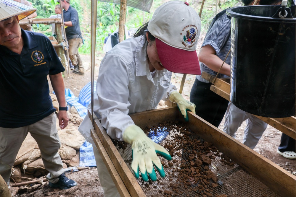 The image shows what appears to be a field excavation or archaeological recovery operation in a forested, tropical setting. A person wearing a white shirt, baseball cap with a U.S. government emblem, and green work gloves is carefully sifting through soil and debris on a wooden-framed mesh screen. The setting and insignia suggest this may be a DPAA (Defense POW/MIA Accounting Agency) or similar U.S. government recovery mission, likely in Southeast Asia.