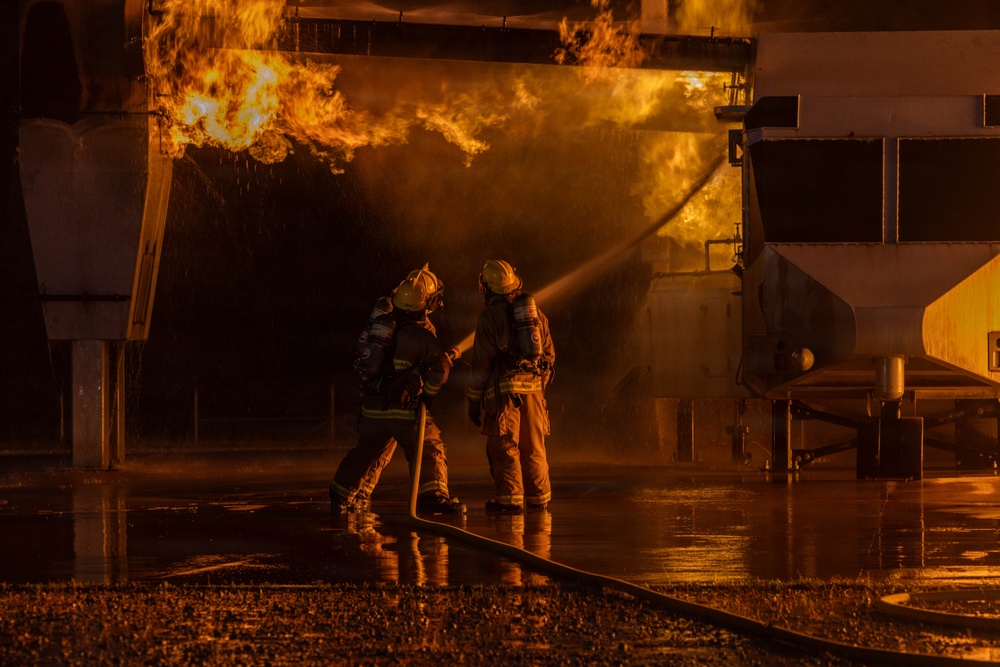 Two firefighters in full protective gear are actively combating a large industrial fire at night. They are operating a fire hose together, directing water spray toward intense flames engulfing what appears to be an industrial structure or vehicle. The wet ground reflects the orange glow of the fire, indicating significant water has already been deployed.