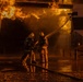 Two firefighters in full protective gear are actively combating a large industrial fire at night. They are operating a fire hose together, directing water spray toward intense flames engulfing what appears to be an industrial structure or vehicle. The wet ground reflects the orange glow of the fire, indicating significant water has already been deployed.