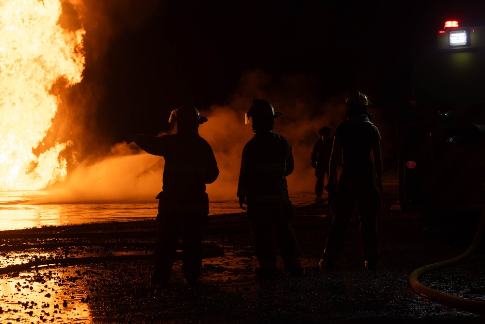 A nighttime firefighting operation showing multiple firefighters in silhouette facing a large, intense fire. The scene is dramatically lit by the orange glow of the blaze, with visible smoke, water spray, and fire hoses on the ground. A vehicle with active lights is visible in the upper right background.