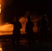 A nighttime firefighting operation showing multiple firefighters in silhouette facing a large, intense fire. The scene is dramatically lit by the orange glow of the blaze, with visible smoke, water spray, and fire hoses on the ground. A vehicle with active lights is visible in the upper right background.