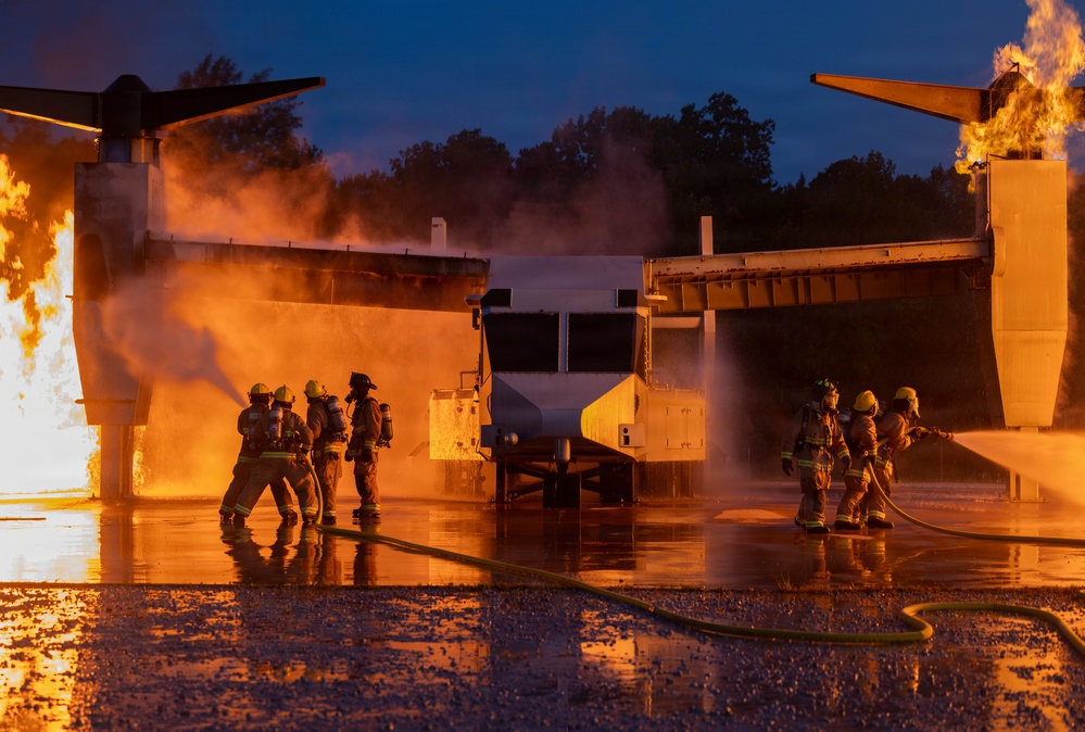 Firefighters in full protective gear and SCBA equipment conduct a nighttime aircraft rescue and firefighting (ARFF) training exercise. The training prop appears to be a mock-up of a V-22 Osprey tiltrotor aircraft engulfed in controlled flames at both engine nacelles. Water and foam suppression efforts are visible from multiple hose teams on both sides of the aircraft.