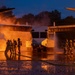 Firefighters in full protective gear and SCBA equipment conduct a nighttime aircraft rescue and firefighting (ARFF) training exercise. The training prop appears to be a mock-up of a V-22 Osprey tiltrotor aircraft engulfed in controlled flames at both engine nacelles. Water and foam suppression efforts are visible from multiple hose teams on both sides of the aircraft.