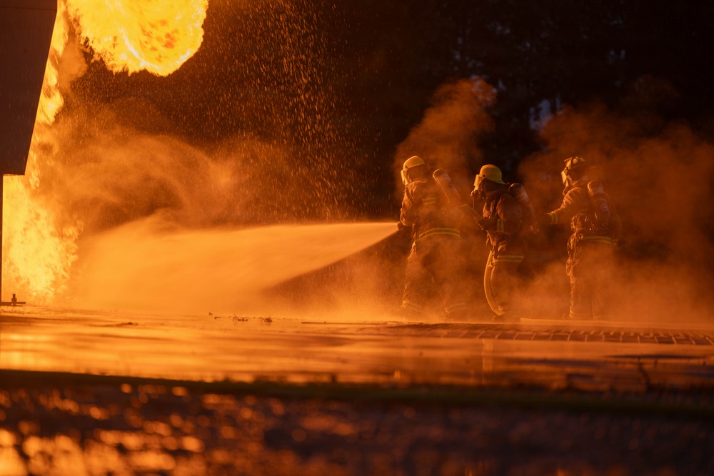 A group of firefighters in full protective gear are actively battling a large, intense fire at night. They are directing a high-pressure water hose stream toward the blaze, which is producing massive orange flames and thick smoke. The scene is dramatically lit entirely by the orange glow of the fire.