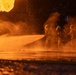 A group of firefighters in full protective gear are actively battling a large, intense fire at night. They are directing a high-pressure water hose stream toward the blaze, which is producing massive orange flames and thick smoke. The scene is dramatically lit entirely by the orange glow of the fire.
