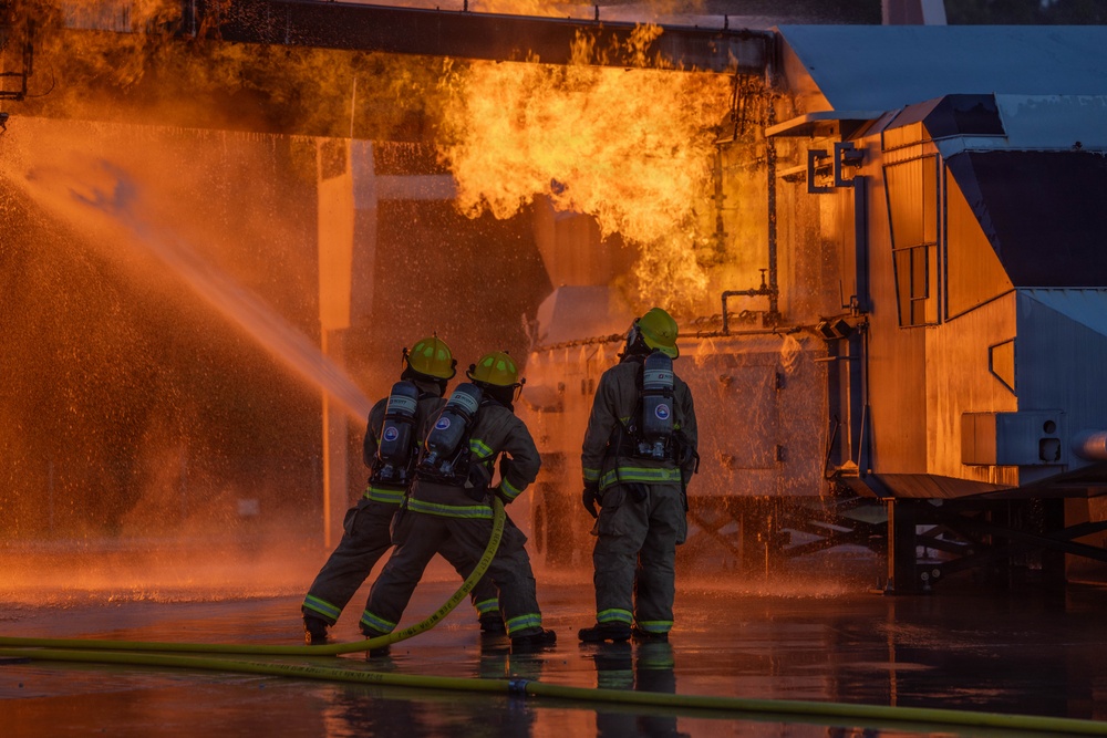 Three firefighters in full protective gear are actively combating a large industrial fire using a hose line. The scene shows intense orange flames erupting from what appears to be an industrial structure or training facility. The ground is wet from water application, and the image is dramatically lit by the fire's glow.