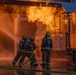 Three firefighters in full protective gear are actively combating a large industrial fire using a hose line. The scene shows intense orange flames erupting from what appears to be an industrial structure or training facility. The ground is wet from water application, and the image is dramatically lit by the fire's glow.