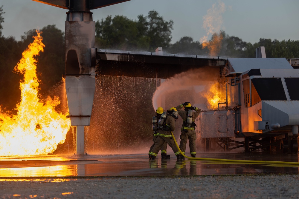Two firefighters in full protective gear are actively combating a large fire using a yellow hose, directing a powerful water stream toward the flames. The scene appears to be a firefighting training exercise involving a mock aircraft or industrial structure with controlled gas-fed fires on multiple sides. The wet ground and structured burn props suggest a controlled training environment.