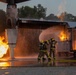 Two firefighters in full protective gear are actively combating a large fire using a yellow hose, directing a powerful water stream toward the flames. The scene appears to be a firefighting training exercise involving a mock aircraft or industrial structure with controlled gas-fed fires on multiple sides. The wet ground and structured burn props suggest a controlled training environment.