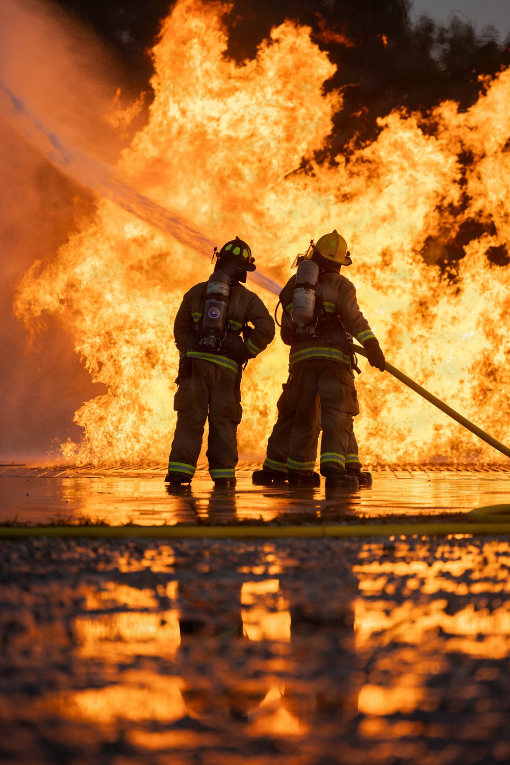 Two firefighters in full personal protective equipment are directing a fire hose toward a massive wall of flames. Both personnel are equipped with SCBA (Self-Contained Breathing Apparatus) tanks and are wearing standard structural firefighting turnout gear with high-visibility reflective striping. The ground is wet and reflective, suggesting significant water has already been deployed, and the fire remains large and active in the background.