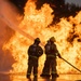 Two firefighters in full personal protective equipment are directing a fire hose toward a massive wall of flames. Both personnel are equipped with SCBA (Self-Contained Breathing Apparatus) tanks and are wearing standard structural firefighting turnout gear with high-visibility reflective striping. The ground is wet and reflective, suggesting significant water has already been deployed, and the fire remains large and active in the background.