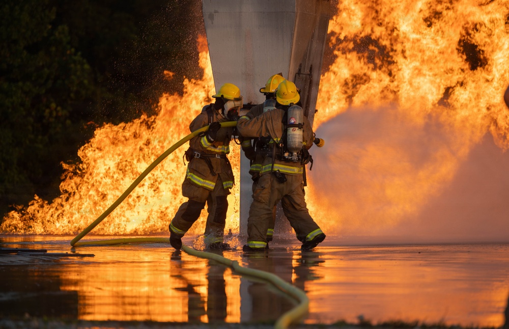 Two firefighters in full protective gear are actively combating a large, intense fire using a yellow hose. They are positioned close together near a structural column, with massive flames visible on both sides. The ground is wet and reflective, indicating significant water has already been deployed.