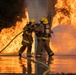 Two firefighters in full protective gear are actively combating a large, intense fire using a yellow hose. They are positioned close together near a structural column, with massive flames visible on both sides. The ground is wet and reflective, indicating significant water has already been deployed.