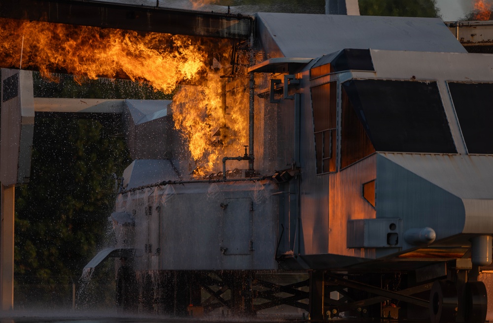 The image shows a large industrial or locomotive vehicle engulfed in significant flames and fire, with visible water spray suggesting active firefighting or suppression efforts. The fire appears to be emanating from the upper mid-section of what appears to be a train locomotive or heavy industrial equipment. The scene is captured in sharp detail with dramatic orange and yellow flames contrasting against the metallic gray structure.
