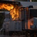 The image shows a large industrial or locomotive vehicle engulfed in significant flames and fire, with visible water spray suggesting active firefighting or suppression efforts. The fire appears to be emanating from the upper mid-section of what appears to be a train locomotive or heavy industrial equipment. The scene is captured in sharp detail with dramatic orange and yellow flames contrasting against the metallic gray structure.
