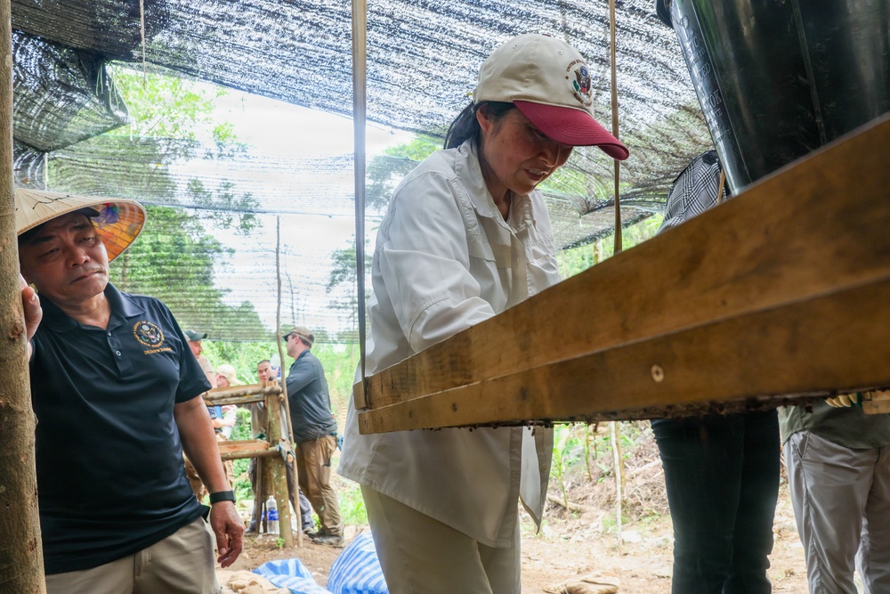 A group of people are gathered under a shaded outdoor structure, likely at a field site or excavation area. A woman wearing a white shirt and a cream-colored cap with a red brim is examining or operating a wooden sifting or screening apparatus. Several other individuals are visible in the background, suggesting a supervised or organized field activity.