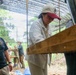 A group of people are gathered under a shaded outdoor structure, likely at a field site or excavation area. A woman wearing a white shirt and a cream-colored cap with a red brim is examining or operating a wooden sifting or screening apparatus. Several other individuals are visible in the background, suggesting a supervised or organized field activity.
