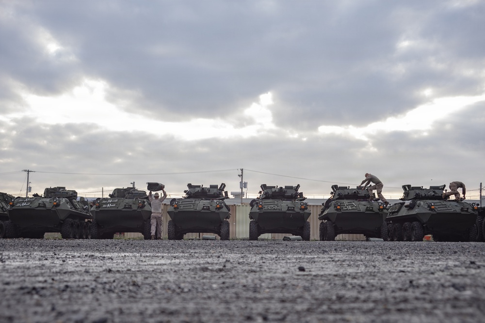 A row of military wheeled armored vehicles, consistent with Light Armored Vehicles (LAVs), are parked in a staging area on a wet, dark gravel surface under an overcast sky. Several personnel in military-style uniforms are performing maintenance or preparation tasks on and around the vehicles. The scene suggests a pre-deployment or post-arrival equipment inspection and servicing operation.