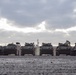 A row of military wheeled armored vehicles, consistent with Light Armored Vehicles (LAVs), are parked in a staging area on a wet, dark gravel surface under an overcast sky. Several personnel in military-style uniforms are performing maintenance or preparation tasks on and around the vehicles. The scene suggests a pre-deployment or post-arrival equipment inspection and servicing operation.