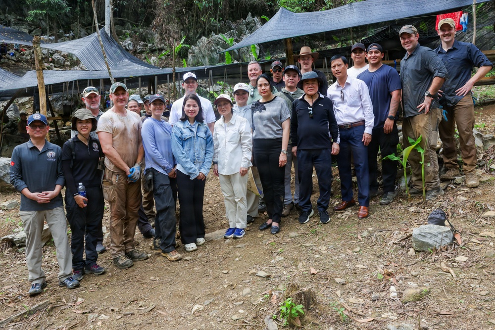 A large group of approximately 20 people pose together for a group photograph at what appears to be an outdoor field site or excavation area in a forested, rocky terrain. The group appears to be a mixed team of civilians and personnel in field attire, gathered under a shade structure made of dark netting. The setting suggests a collaborative field operation, possibly archaeological, humanitarian, or recovery-related work.