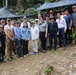 A large group of approximately 20 people pose together for a group photograph at what appears to be an outdoor field site or excavation area in a forested, rocky terrain. The group appears to be a mixed team of civilians and personnel in field attire, gathered under a shade structure made of dark netting. The setting suggests a collaborative field operation, possibly archaeological, humanitarian, or recovery-related work.
