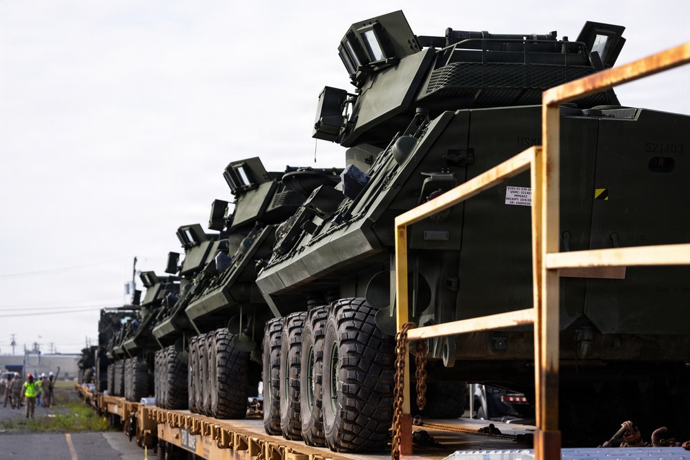 Multiple military wheeled armored vehicles are loaded on flatbed rail cars or trailers, secured with chains, appearing to be in a transport or deployment configuration. The vehicles are painted in standard military olive drab/dark green and appear to be modern armored fighting vehicles, possibly M-ATV or similar MRAP-class vehicles. Personnel in high-visibility vests are visible in the background, suggesting an active loading or staging operation.