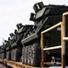 Multiple military wheeled armored vehicles are loaded on flatbed rail cars or trailers, secured with chains, appearing to be in a transport or deployment configuration. The vehicles are painted in standard military olive drab/dark green and appear to be modern armored fighting vehicles, possibly M-ATV or similar MRAP-class vehicles. Personnel in high-visibility vests are visible in the background, suggesting an active loading or staging operation.