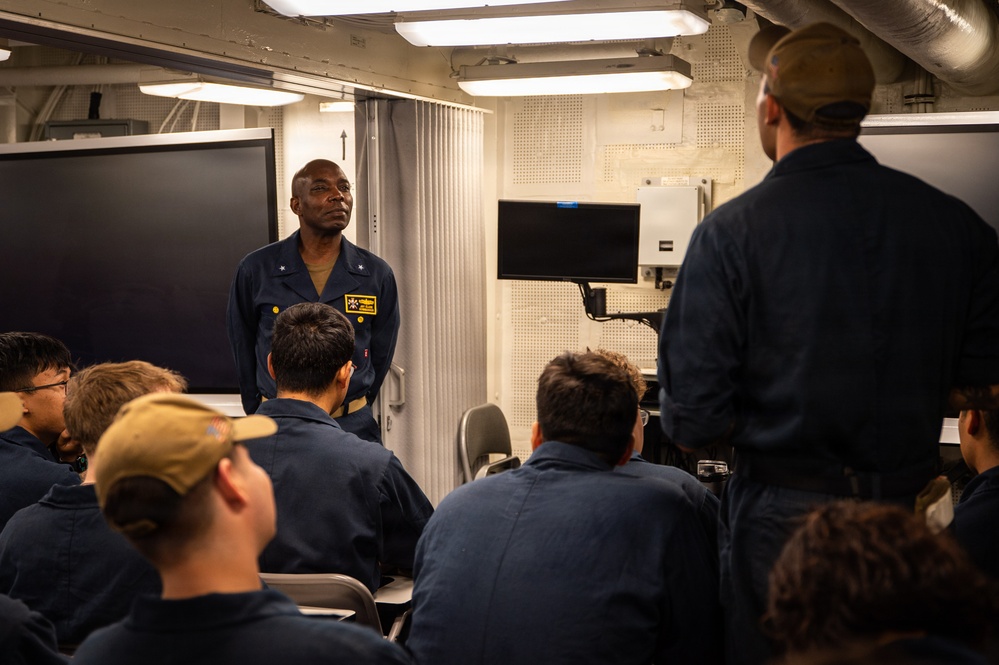 A military briefing or training session is taking place in what appears to be a shipboard classroom or briefing room. A senior naval officer or petty officer in dark blue coveralls stands at the front addressing a group of seated personnel, also in dark blue coveralls. The setting, lighting, and equipment are consistent with a U.S. Navy vessel interior.