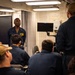 A military briefing or training session is taking place in what appears to be a shipboard classroom or briefing room. A senior naval officer or petty officer in dark blue coveralls stands at the front addressing a group of seated personnel, also in dark blue coveralls. The setting, lighting, and equipment are consistent with a U.S. Navy vessel interior.