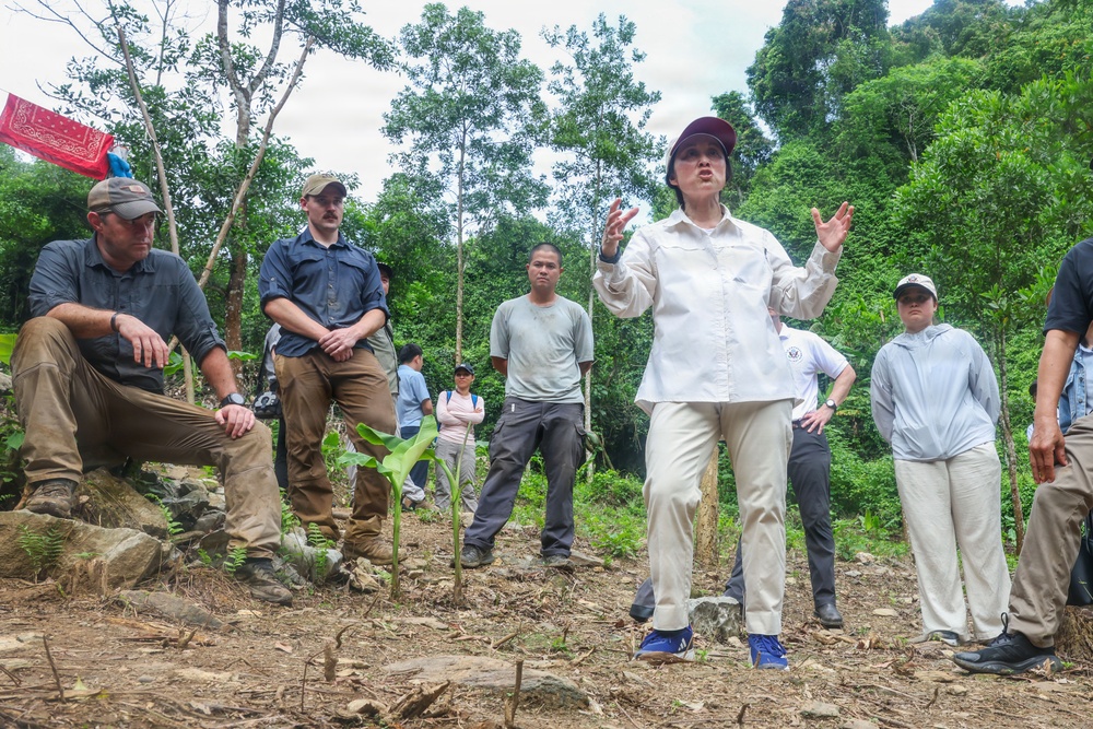 A group of approximately 8-10 people are gathered in a forested, hilly outdoor setting. A woman in a white long-sleeve shirt, khaki pants, and a dark cap appears to be leading a briefing or explanation, gesturing with her hands while others listen attentively. The setting suggests a field survey, archaeological dig, or environmental assessment mission.