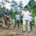 A group of approximately 8-10 people are gathered in a forested, hilly outdoor setting. A woman in a white long-sleeve shirt, khaki pants, and a dark cap appears to be leading a briefing or explanation, gesturing with her hands while others listen attentively. The setting suggests a field survey, archaeological dig, or environmental assessment mission.