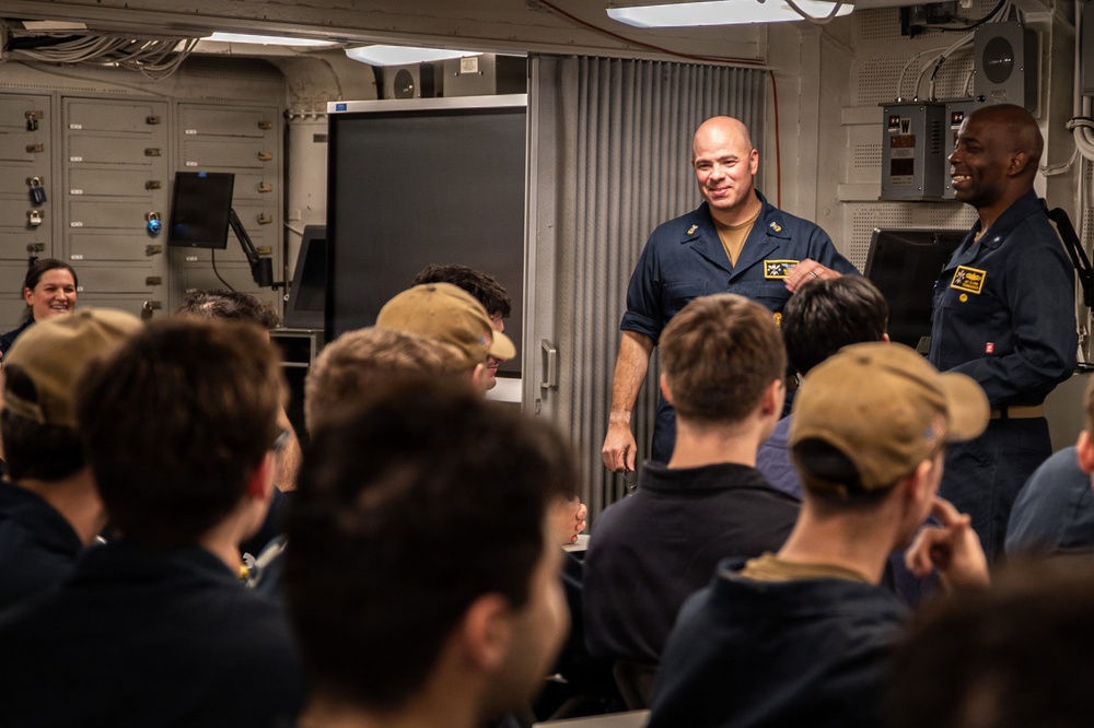 A briefing or meeting is taking place in what appears to be a military vessel's interior compartment. Two uniformed personnel, likely U.S. Navy sailors based on their dark blue coveralls and insignia patches, are standing and addressing a seated group of individuals. The setting has the characteristic appearance of a shipboard briefing room with metal walls, lockers, and a large display screen.